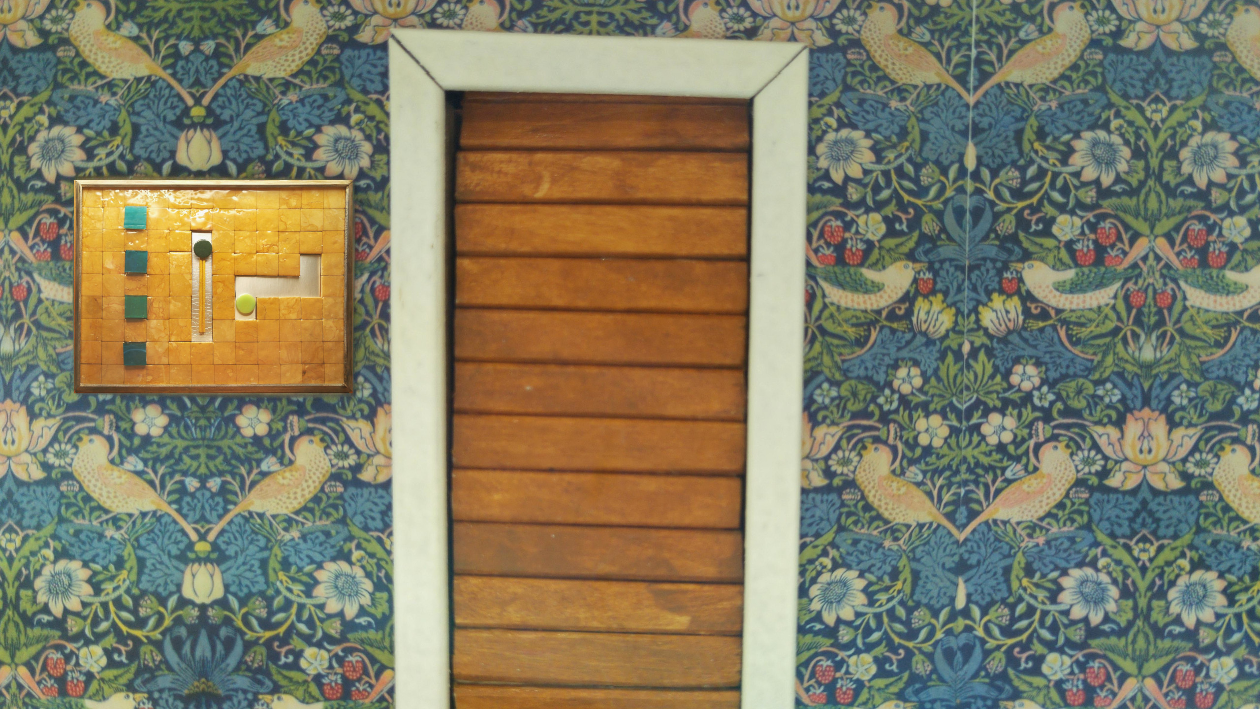 In a wall papered with William Morris wallpaper, we see a doorway with a door that is intended to slide up.  To the left, a mosaic is mounted on the wall.  It has four buttons and two tracks on which circular tiles might slide.